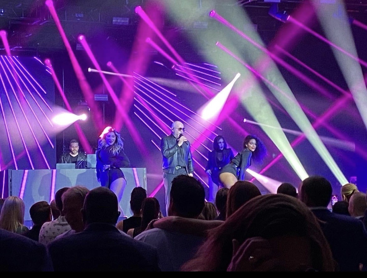 Live stage performance with singer, dancers, and DJ illuminated by vibrant purple and white intelligent lighting beams.