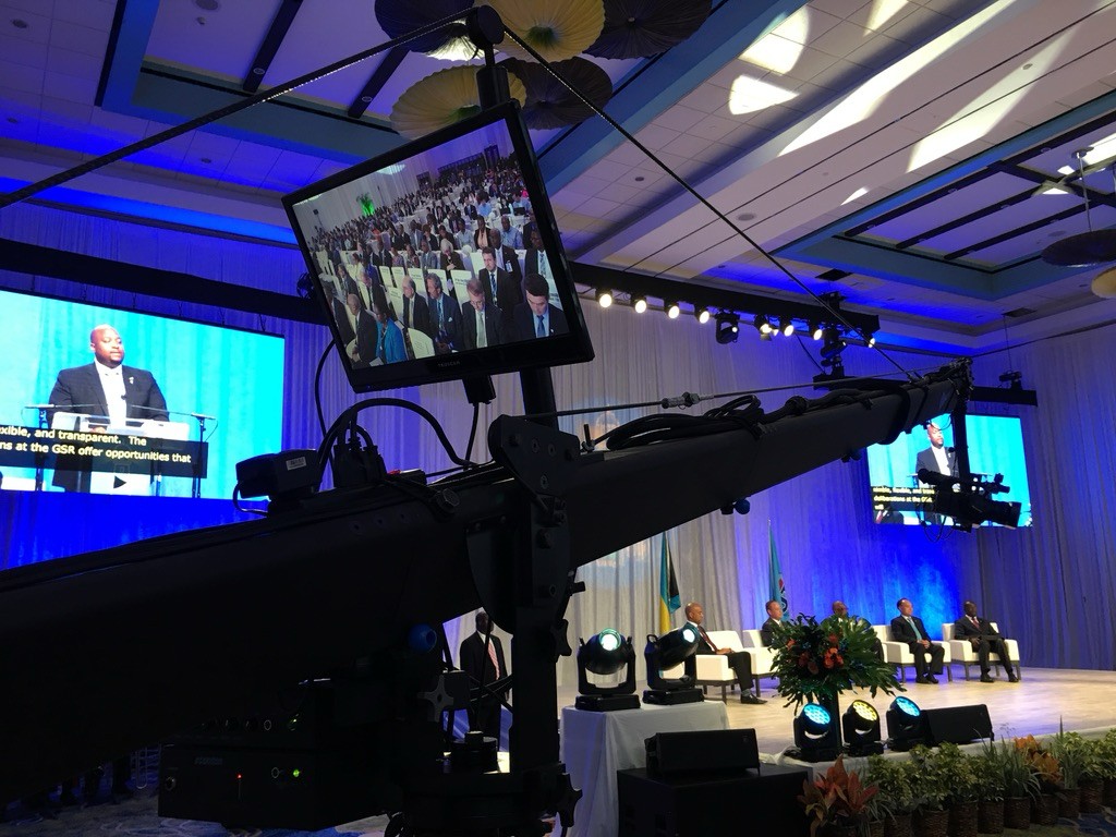 Camera crane filming a conference stage with speakers and a large presentation screen.